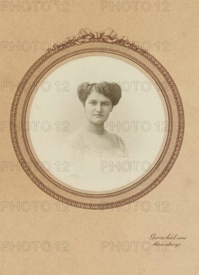Passepartout of Mathilde Wachenheimer-Wertheimer as a young woman, photograph, in light clothing., photographer: Gerschel aîné, Strasbourg (France), 1880 - 1895, paper, gelatin silver print, developed gelatin silver print, height 165 mm x width 105 mm, Person, Mathilde Wachenheimer-Wertheimer, Period, A sepia-toned portrait of a woman is centered within an ornate oval frame printed on textured paper; the woman faces forward with a neutral expression, her dark hair styled in a symmetrical updo, and she wears a light-colored dress with lace trim and a delicate necklace, while the decorative oval border features intricate patterned bands and a ribbon-like motif at the top.