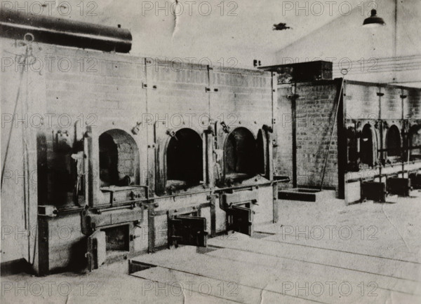 Incinerators in a concentration camp, photograph, Suspected incinerators from an unknown concentration camp., 1940 - 1945, photographer, paper, gelatin silver print, developed gelatin silver print, height 90 mm x width 115 mm, Period, A black and white photograph showing a long row of brick furnaces or ovens with arched openings set into a tall masonry wall, each opening framed by metal doors and hinges, with some lower metal access panels open and rails or tracks running along the front; a platform or catwalk with support structures is visible above some of the ovens, and a hanging industrial light fixture appears near the ceiling; pipes, metal brackets, cables, and pulleys run along the wall and ceiling, and the foreground shows a smooth floor with faint lines and a recessed rectangular opening.