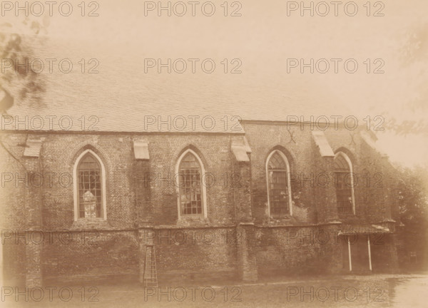 South side of the St. Nicholas Church in Dwingeloo, photograph, annotation: '12', annotation: 'Dwingelo', Dwingeloo, 1890 - 1910, photographer, Attributed to, paper, pasteboard, albumen print, height 166 mm x width 228 mm, church (exterior), Sint-Nicolaaskerk (Dwingeloo), A sepia-toned photograph of a brick building wall with a sloped tiled roof, showing a row of tall arched windows with multi-pane glazing set into the brickwork, buttresses between the window bays, small stone ledges above some sections, a wooden ladder leaning against the wall, tree branches and leaves partially visible at the image edges, and a ground area in front of the building.