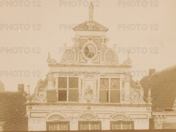 Facade of Friesestraat 9 in Coevorden, photograph, annotation: 'Friesesraat D.489, Coevorden', annotation: 'I', Coevorden, 1890 - 1910, photographer, Attributed to, paper, pasteboard, albumen print, albumendruk, height 168 mm x width 225 mm, façade (of house or building), An ornate building facade with decorative scrollwork and sculpted figures, a triangular pediment topped by a standing statue, an oval window with radial muntins centered beneath the pediment, several rectangular windows with diamond-pane glazing and open shutters, carved ornamental panels and leafy motifs around the openings, curved rooftop elements and finials on either side, a pair of stylized shell-shaped reliefs above lower windows, portions of sloped tiled roofs visible at the sides, and a muted sepia tone across the entire image.