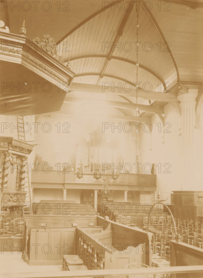 Interior of the Reformed Church in Coevorden, photograph, annotation: 'IV', annotation: 'Church in Coevorden', Coevorden, 1890 - 1910, photographer, Attributed to, paper, pasteboard, albumen print, height 229 mm x width 165 mm, interior of church, A sepia-toned interior view showing wooden pews arranged in rows, carved railings and paneling, ornate decorative woodwork on a raised pulpit with spiral columns, a balcony with wooden front and columns beneath it, a suspended chandelier and a vertical hanging chain of light fixtures, a vaulted wooden ceiling with curved beams, a tall fluted column against the right wall, and various chairs and wooden furniture scattered throughout the space.
