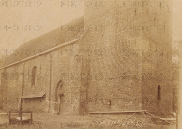 Northwest side of the Reformed Church in Westerwijtwerd, photograph, annotation: 'Westerwijtwerd, municipality of Middelstum', annotation: 'Church in Westerwijtwerd, NW view July 1897, Westerwijtwerd, 1897, photographer, Attributed to, paper, pasteboard, albumen print, height 161 mm x width 218 mm, church (exterior), A large brick building with a steep tiled roof and a tall tower section fills the frame, showing textured masonry, narrow arched windows, and vertical metal rods attached to the walls; a small doorway set into an arched recess and a covered wooden lean-to are visible along one wall, and in front of the building there is a wooden crate-like structure on grass, scattered stones, and a low wooden frame partially covering a shallow brick-lined pit.