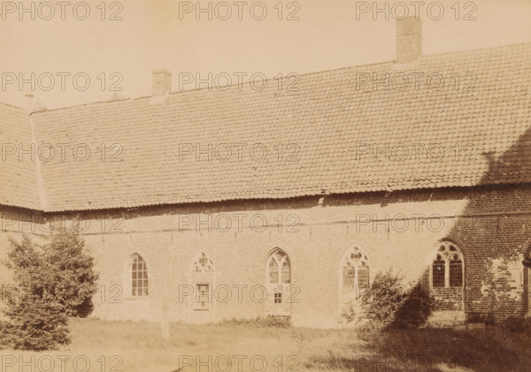 Exterior of the cloister of Ter Apel Monastery, photograph, annotation: 'Ter Apel', annotation: 'Ter Apel, Cloister wall east of the courtyard, Sept 1893', Ter Apel, 1893, photographer, Attributed to, paper, pasteboard, albumen print, height 168 mm x width 232 mm, courtyard, cloisters, monastery, A large brick building with a steep tiled roof and two visible chimneys occupies most of the image, showing a row of arched windows with decorative tracery along the upper wall and rectangular windows and a door below, with a grassy area and several shrubs and trees in the foreground and a shadow falling across part of the roof and wall.