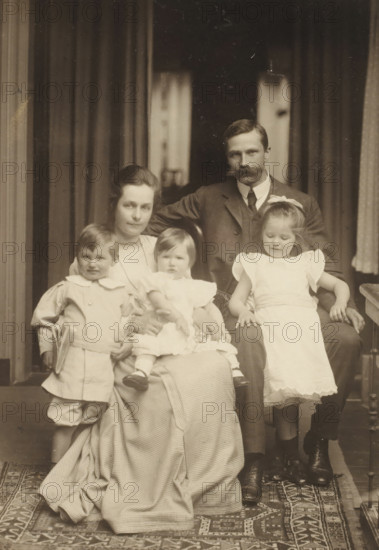 Family portrait of the Van der Waals family, photograph, annotation: 'Of Cor, Hansje, Jan, Anke, June 1907. Groningen', photographer: Aleidas Christiaan Kunst, Groningen (city), 1907, paper, pasteboard, daylight collodion silver print, height 231 mm x width 169 mm, Person, Jennigje Hendrika Jacoba Jonker, Johannes Diderik van der Waals (1873-1971), Johanna Geertruida van der Waals, Anne Madeleine van der Waals, historical persons, anonymous historical persons portrayed in a group, in a group-portrait, A sepia-toned portrait of two adults and three children seated and standing on a patterned rug in front of heavy curtains, with the man positioned behind and slightly to the side of the woman, wearing a dark suit and sporting a mustache, and the woman wearing a light-colored blouse and a long checked skirt while holding a small child in a white dress on her lap; a young boy in a light-colored sailor-style outfit stands to one side holding a book, and a young girl in a white dress with puf...
