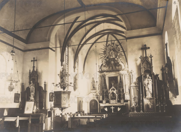 Interior of the former Roman Catholic church in Horssen, photograph, annotation: 'B', annotation: 'Horssen (G.), Oct 1908', Horssen, 1908, photographer, Attributed to, paper, pasteboard, gelatin silver print, height 160 mm x width 217 mm, interior of church, The image shows the interior of a church with vaulted ceilings and arched ribs, chandeliers and hanging lamps suspended from the ceiling, stained glass windows along the side walls, wooden pews in rows facing an ornate altar area, multiple statues of religious figures placed on pedestals and in niches, tall decorative crosses atop wooden altarpieces, an elaborate central altarpiece with radiating decorative elements behind a statue, framed artwork and candlesticks on stands, and carved wooden furnishings and panels throughout the space.