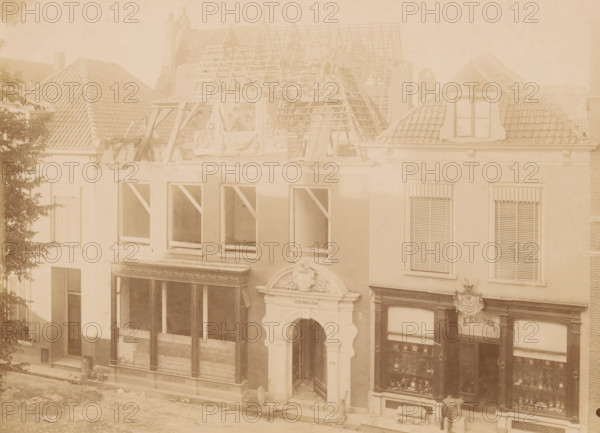 View of Kleine Poot 9 in Deventer, photograph, annotation: 'Deventer, Huis Kleine Poot 9, demolished July 1901', Deventer, c. 1901, photographer, Attributed to, paper, pasteboard, albumen print, albumendruk, height 165 mm x width 229 mm, urban housing, street (city(-scape) with figures, staffage), façade (of house or building), A faded sepia view of a row of adjacent buildings with tiled roofs, dormer windows, and scaffolding on one roof where wooden beams and ladders are visible and a few indistinct figures sit or stand; the façades show multiple window openings with some frames boarded or supported by diagonal braces, decorative stone and plaster ornamentation above a central arched entrance, storefront windows with display cases and decorative moldings, a person standing near one shop entrance, and a tree partially obscuring the left edge of the scene.