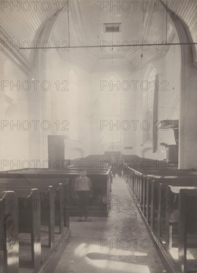 Interior of the Reformed Church in Hellendoorn, photograph, annotation: 'Hellendoorn, Dutch Reformed Church, Sept 1909', Hellendoorn, 1909, photographer, Attributed to, paper, pasteboard, gelatin silver print, height 222 mm x width 163 mm, interior of church, A high, vaulted interior space with wooden pews arranged in rows on either side of a central aisle, tall arched windows along the far wall admitting diffuse light, a curved wooden ceiling with visible paneling and suspended fixtures, a raised pulpit or lectern projecting from the right wall, narrow walkways between pews, and a group of small, blurred figures standing near the back beneath the large windows.