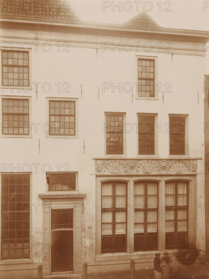 Façade of the Buiten-Proveniershuis in Kampen, photograph, annotation: 'Kampen, The Buiten-Proveniershuis on Nieuwstraat, Nov 1896', annotation: 'Kampen', Kampen, 1896, photographer, Attributed to, paper, pasteboard, daylight collodion silver print, height 227 mm x width 171 mm, urban housing, façade (of house or building), A sepia-toned facade of a multi-story building with rectangular framed windows, some with small panes and others with larger panes and curtains, a decorative stone band with carved fan-shaped motifs above a row of tall windows, a doorway with a lintel and flanked pilasters, smooth plastered walls with visible metal anchors, a tiled roof peeking above the cornice, and blurred silhouettes of two people standing on the sidewalk in front of the building.