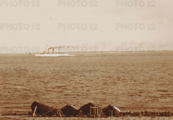 Navy ship Hr. Ms. Zeeland off the coast of Den Helder, 'Zeeland', cabinet photograph, collector's mark: 'Vosmaer', number: '0.50', photographer: Anna Clasina Leijer, Den Helder, 1890 - 1910, Anna Clasina Leijer, photographer, paper, pasteboard, baryta paper, daylight collodion silver print, height 105 mm x width 165 mm, navy, steamship, motorship, sea (seascape), North Sea, A wide view of a calm sea with rippling water filling most of the frame and a pale sky above, a long, low ship steaming along the horizon leaving a trail of dark smoke, several small sail or marker shapes visible near the distant shoreline, and a row of small wooden boats pulled up on a rocky shore in the foreground with ropes and planks nearby.