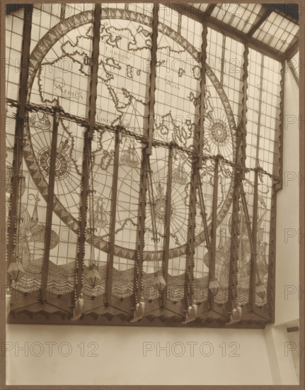 Stained glass window with continents and sailing ships, photograph, check stamp: 'BERN.P.EILERS, AMSTER, DAM', photographer: Bernard Eilers, 1888 - 1951, paper, daylight collodion silver print, height 220 mm x width 170 mm, work of monumental art, maps of the continents and the polar regions, A large decorative window fills the upper portion of the image, composed of many rectangular panes and divided by vertical dark beams; the window contains an intricate circular map-like design with ornate compass roses, stylized coastlines, and sailing ships, rendered in fine linework across the panes. Decorative elements hang from the top of the frame, including patterned bands and suspended geometric lantern shapes, with sculpted figures positioned along the lower edge of the window framework. The lower portion of the photograph shows a plain light-colored wall beneath the window and portions of adjacent architectural surfaces framing the scene. The image has a monochromatic, sepia-toned appeara...