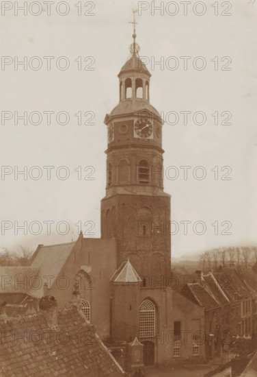 Tower of the St. Lambertus Church in Buren, photograph, annotation: 'NH Kerk, Buren', annotation: 'Tower of the Dutch Reformed congregation in Buren (St. Lambertus)', Buren, 1890 - 1920, photographer, paper, pasteboard, daylight collodion silver print, height 424 mm x width 295 mm, parts of church exterior and annexes: tower, A tall brick church tower rises above surrounding rooftops, topped with a multi-tiered belfry and a domed roof with open arched openings; a clock face is visible on the upper section of the tower, and circular decorative elements flank it; the tower shows arched brickwork and rectangular louvered openings below the clock, and a small conical turret projects from the side of the lower tower above a large arched window with a grid of panes; steep tiled roofs of adjacent buildings and a ridge of rooftops frame the foreground, and rows of bare trees and distant low buildings are visible along the horizon beneath a pale cloudy sky.