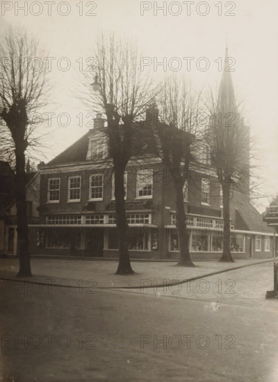 View of Spruyt van Rietschoten shop in Hilversum, photograph, inscription: 'Vosmaer', annotation: 'Hilversum', Hilversum, 1900 - 1930, photographer, paper, gelatin silver print, developed gelatin silver print, height 180 mm x width 130 mm, shop, store, A street scene showing a two-story brick building with a tiled roof and large display windows at ground level, flanked by leafless trees whose bare branches rise in front of the façade and partially obscure the upper windows and roofline, with a tall pointed church spire visible in the background behind the trees, a sidewalk and curb in front of the building, and a paved roadway in the foreground.