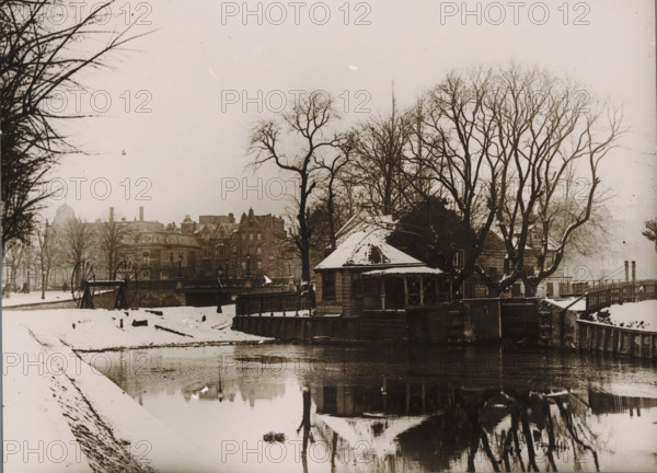 View of a small house on the Stadhouderskade in Amsterdam, photograph, annotation: 'Rijksprentenkabinet, received on loan from mus. Amstelkring, April 1952', annotation: 'Received 10 Nov 1948, glass broken', annotation: 'Small house Stadhouderskade near Rijksmuseum', annotation: '...', Amsterdam, 1900 - 1930, photographer, paper, gelatin silver print, developed gelatin silver print, height 172 mm x width 236 mm, urban housing, street, A snowy riverside scene showing a small wooden building with a pitched roof partially covered in snow, positioned at the water’s edge and surrounded by fencing and wooden posts, with bare trees rising behind and around it. The river reflects the building and trees, and a snow-covered bank slopes down to the water in the foreground. Across the river a bridge or embankment is visible with large spoked wheels mounted on stands nearby, and behind that a row of multi-story stone buildings with chimneys and architectural detailing extends along the skyline. Lea...