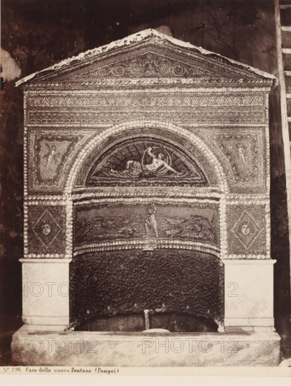 Mosaic in the Casa della nuova Fontana in Pompeii, Italy, Casa della nuova fontana (Pompei), photograph, number: '21', collector's mark: 'RICH SCHOOL FOR ART HANDBOOK, GROUP', number: 'No. 1261.', photographer: Giorgio Sommer, Pompeii, 1857 - 1875, Giorgio Sommer, photographer, pasteboard, paper, albumen print, height 478 mm x width 319 mm, urban housing, ornamental fountain, work of monumental art, Casa della Nuova Fontana, A decorative architectural niche with an ornate triangular pediment sits above an arched recess, the surface covered in dense mosaic patterns and small rounded accents. Inside the arched lunette a reclining figure is depicted within a circular frame, and panels flanking the arch contain smaller human figures. The vertical sides show diamond-shaped mosaic panels with circular motifs, and the lower area beneath the arch is filled with a rough, dark textured surface leading down to a stone basin supported by plain rectangular pilasters. A narrow ledge runs along the b...