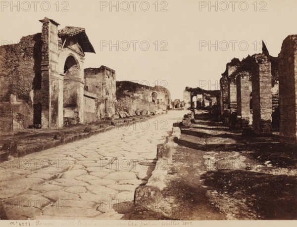 View of the Porta Ercolano in Pompeii, Italy, Porte Erculaneum faction fouillés 1812, Pompei, photograph, collector's mark: 'VOSMAER', number: 'No. 4221', photographer: attributed to Giorgio Sommer, Pompeii, 1857 - 1875, Giorgio Sommer, photographer, Attributed to, pasteboard, paper, albumen print, height 308 mm x width 405 mm, street, ruin of a building, architecture, landscape with ruins, gate, entrance, Porta Ercolano, A wide ancient street paved with large irregular stones runs from the foreground toward the background, flanked on the left by weathered stone and brick ruins including a tall arched doorway topped by a tiled roof and framed by textured walls, and on the right by rows of ruined stone columns and wall fragments casting long shadows on uneven ground, with scattered stone blocks and remnants of masonry along both sides and larger archways visible further down the street beneath a pale empty sky.