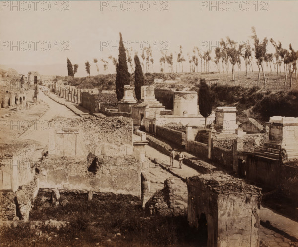 View of the Strada delle Tombe in Pompeii, Italy, Strada delle Tombe, Pompei, photograph, collector's mark: 'VOSMAER', number: '1278', photographer: Giorgio Sommer, Pompeii, 1857 - 1875, Giorgio Sommer, photographer, pasteboard, paper, albumen print, height 315 mm x width 422 mm, street, ruin of a building, architecture, landscape with ruins, gravestone, tombstone, cemetery, park-like burying ground, Via delle Tombe (Pompeï), A wide view of an ancient ruined street flanked by crumbling stone walls and partially standing structures, with paved pathway running through the center, scattered rectangular tomb-like monuments and raised stone platforms along one side, tall narrow cypress trees and a line of thinner trees visible on the ridge beyond the ruins, grassy patches and rubble filling open areas, two figures walking on the paved path, and the scene rendered in a sepia-toned photographic style.