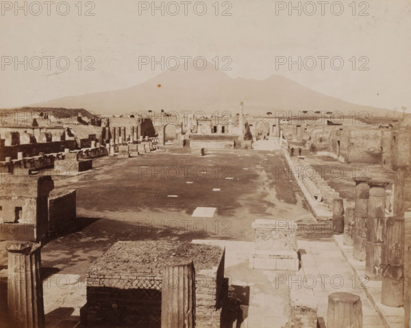 View of the Forum in Pompeii, Pompei, photograph, number: '1202.', collector's mark: 'VOSMAER', photographer: attributed to Giorgio Sommer, Pompeii, 1863 - 1875, Giorgio Sommer, photographer, Attributed to, pasteboard, paper, albumen print, height 314 mm x width 421 mm, In the background right of the square another photographer is at work., portrait, self-portrait of photographer, square, circus, ruin of a building, architecture, landscape with ruins, Piazza del Foro, A wide sepia-toned view of ancient ruins with rows of stone columns, broken walls, and rectangular stone blocks stretching into the distance, an open paved central area with scattered square stones, a raised platform and archways toward the middle ground, two small figures standing near a paved walkway at the far right, low vegetation and a line of trees visible beyond the ruins, and a large gently sloping mountain forming the background under a pale sky.