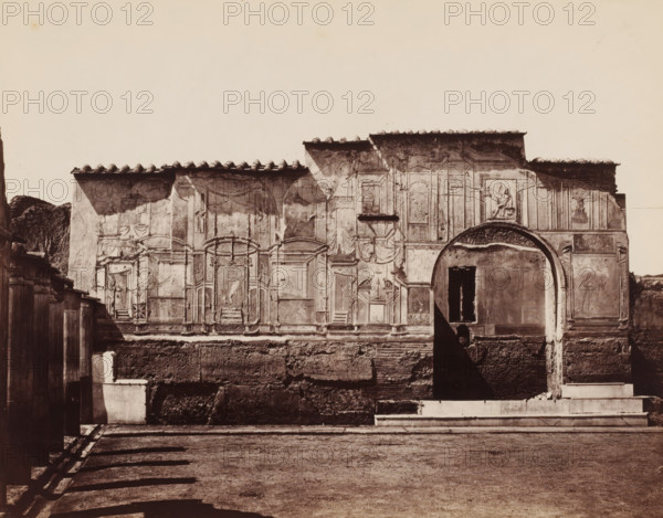 Bagni nuovi (Pompeii), Ruins of a bathhouse in Pompeii, photograph, number: '28', inscription: 'Pompei', number: '29', collector's mark: 'RICH SCHOOL FOR ARTS HANDBOOK, GROUP', number: 'No. 1258.', photographer: attributed to Giorgio Sommer, Pompeii, 1863 - 1875, Giorgio Sommer, photographer, Attributed to, pasteboard, paper, albumen print, height 320 mm x width 478 mm, ruin of a building, architecture, landscape with ruins, public baths, Bagni nuovi, A sunlit facade of an old stone building fills the middle of the image, showing faded decorative panels and reliefs, a large arched opening with a dark interior, and a rectangular window above a worn wall; tiled roof eaves cast shadows along the top edge and small sculpted figures appear near the arch; the lower portion of the wall shows exposed brick and rough texture, with a stepped platform and a shallow paved foreground in front; on the left side a wooden fence and a small plastered structure are visible, and long diagonal shadows fal...