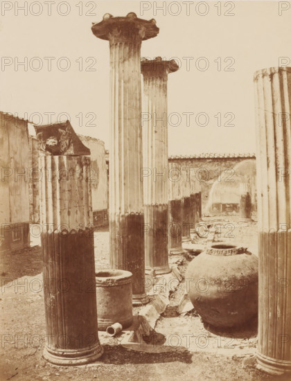 Ruin in Pompeii, Italy, photograph, collector's mark: 'VOSMAER', photographer: attributed to Giorgio Sommer, Pompeii, 1857 - 1875, Giorgio Sommer, photographer, Attributed to, pasteboard, paper, albumen print, albumendruk, height 405 mm x width 306 mm, urban housing, ruin of a building, architecture, column, pillar, A sepia-toned photograph showing a row of tall, fluted stone columns with ornate capitals, some standing intact and others partially broken, set along a paved trough or channel with fragments and cylindrical stone bases scattered at ground level; in the foreground a large rounded amphora-like clay jar sits beside the channel, and smaller stone vessels and a short hollow drum are visible near the columns; low ruined walls and a tiled roofline run along the background, with distant columns and wall surfaces showing worn and cracked textures.