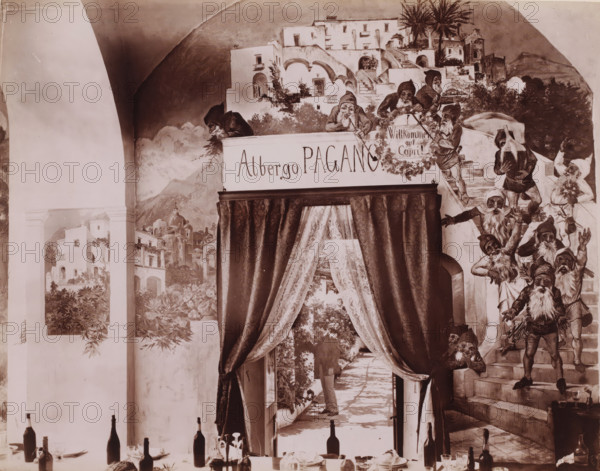 Interior of the dining room of Hotel Pagano on Capri, Italy, Capri, photograph, inscription: 'Capri', number: '6125', photographer: attributed to Giorgio Sommer, 1857 - 1914, Attributed to, pasteboard, paper, albumen print, height 317 mm x width 445 mm, hotel, hostelry, inn, Isola di, A painted wall scene with architectural elements showing arched openings, stairways, terraces, and clustered white buildings surrounded by foliage, palm trees, and distant hills, populated by many small bearded figures wearing pointed hats and tunics, some standing on steps and others leaning over balconies, a heavy dark curtain with patterned drapery framing a passageway that leads to a sunlit courtyard with potted plants and a person partially visible in the distance, a dining setup in the foreground with bottles, glasses, plates, cutlery, and a few decorative items arranged along a table edge.