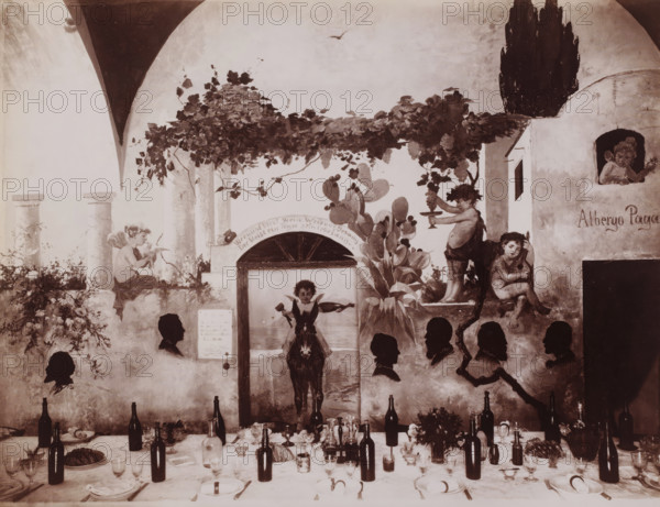 Hotel Pagano, Interior of the dining room of Hotel Pagano in Capri, Italy, Capri, photograph, number: '5170 ?', photographer: attributed to Giorgio Sommer, 1857 - 1914, Attributed to, pasteboard, paper, albumen print, height 318 mm x width 445 mm, hotel, hostelry, inn, Isola di, A painted wall scene shows an archway with climbing vines and leaves overhead, a cactus plant with pads, and sculpted columns. Several cherub-like figures and children are depicted in various poses around the arch and a small building facade, one child holding a platter and another leaning while playing a small instrument. A central painted figure sits astride a donkey visible through an opening beneath the vines. Silhouetted profile heads appear arranged across the lower portion of the wall. In front of the wall a long table is set with plates, folded napkins, glasses, bottles, serving dishes, and floral arrangements, with scattered utensils and a loaf of bread on a platter. The overall palette is muted and se...