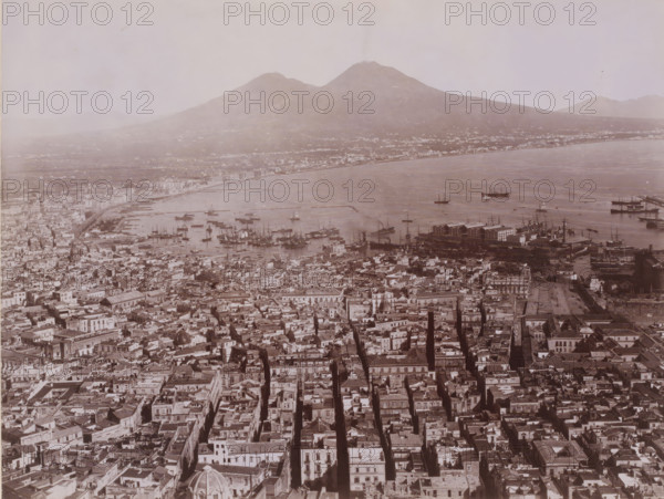 View of Naples, with Mount Vesuvius in the background, Italy, photograph, number: '1103', photographer: Giorgio Sommer (possibly), Naples, 1857 - 1914, pasteboard, paper, albumen print, height 318 mm x width 445 mm, city-view in general, 'veduta', gulf, bay, volcano, Mount Vesuvius, A wide aerial view of a coastal city with densely packed buildings and narrow streets in the foreground, a busy harbor with numerous moored and anchored ships and boats in the midground, and a broad bay stretching toward distant hills and twin-peaked mountains in the background under a pale sky.
