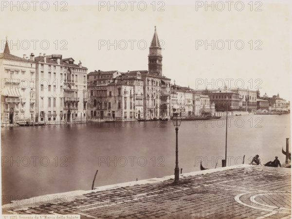 View of the Grand Canal and the Sante Maria della Salute in Venice, Italy, Venezia da Sa Maria della Salute, photograph, address: 'GIORGIO SOMMER, STUDIO MONTE DI DIO 4., MAGAZZINO S. GATERINA 5., NAPOLI', number: 'No. 3525.', photographer: Giorgio Sommer, Venice, 1857 - 1914, pasteboard, paper, albumen print, height 304 mm x width 375 mm, canals, waters (in city), church (exterior), Basilica of Santa Maria della Salute, Grand Canal, A wide waterfront view shows a row of multi-story stone buildings with varied facades and balconies lining a canal, several with awnings and small boats moored at water-level entrances; a tall bell tower rises behind the buildings toward the center of the scene; the foreground features a cobbled promenade with a decorative curved paving pattern, a tall ornate lamppost near the water, and two seated figures at the edge beside a small boat; smooth water occupies the middle ground between the promenade and the buildings.
