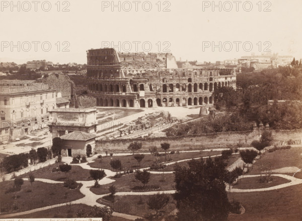 Colosseo preso dai giardini farnesi (Roma), View of the Colosseum in Rome, Italy, photograph, number: '7.', address: 'SOMMER & BEHLES, ROMA E NAPOLI', number: 'No. 1024.', inscription: 'Roma', inscription: 'C. 85', photographer: Sommer & Behles, Rome, 1867 - 1874, pasteboard, paper, albumen print, albumendruk, height 320 mm x width 378 mm, ruin of a building, architecture, amphitheatre, Colosseum, A wide sepia-toned view of an ancient oval stone amphitheater with multiple levels of arched openings, showing weathered and partially ruined exterior walls; in front of it a low rectangular stone structure with a flat top and a building with arched windows and a tiled roof; winding pale pathways curve through a grassy garden area dotted with small trees and shrubs across the foreground; dense trees and taller vegetation occupy the area to the right of the amphitheater and extend into the background; scattered low buildings and ruins appear beyond and to the left of the amphitheater under a p...