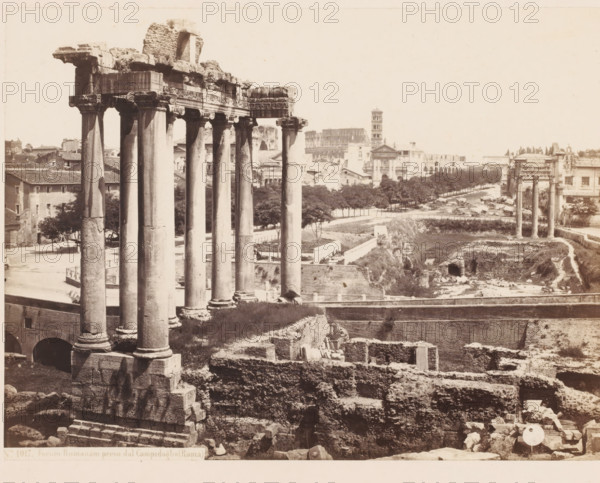 Forum Romanum preso dal Campidoglio (Roma), Ruins at the Roman Forum in Rome, Italy, photograph, inscription: 'Roma, Forum', address: 'SOMMER & BEHLES, ROMA E NAPOLI', number: '4', inscription: 'C. 85', number: 'No. 1017.', photographer: Sommer & Behles, Rome, 1867 - 1874, pasteboard, paper, albumen print, albumendruk, height 319 mm x width 380 mm, square, circus, ruin of a building, architecture, landscape with ruins, Forum Romanum, A wide sepia photograph shows a row of tall ancient stone columns with carved capitals in the foreground standing on large square bases, adjacent to fragmented stone ruins and terraced masonry; behind them a roadway lined with trees leads toward more classical ruins including another set of columns and a distant multi-level stone structure with arches and a tower, while surrounding the scene are low buildings, scattered stones, paths, and patches of grass and exposed earth.