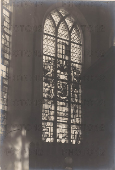 Painted window in the Castle Chapel of Egmond in Egmond aan den Hoef, photograph, annotation: 'stained glass window of Prince Frederik Hendrik, 1634 in the choir of the Dutch Reformed Church. Feb. 1907, Egmond a, d Hoef', annotation: 'Egmond a, d Hoef, stained glass window of Prince Frederik Hendrik 1634 in the choir of the Dutch Reformed Church, 1907', Castle Chapel (Egmond-aan-den-Hoef), photographer: AJM Mulder (possibly), 1907, Egmond aan den Hoef, Attributed to, paper, pasteboard, gelatin silver print, height 218 mm x width 149 mm, interior of church, church-window, e.g. rose-window, A tall arched stained glass window fills the center of the image, its upper sections showing diamond-patterned panes and curved mullions, while the lower panels feature decorative figurative and ornamental glasswork with human figures and heraldic motifs; light filters through the glass casting bright highlights against a dark interior wall, and a narrower stained glass window is partially visible alo...
