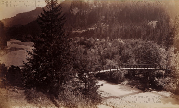 Pont Turrian pres Chateau d'Oex, View of the Pont Turrian in Château-d'Œx, Switzerland, photograph, collector's mark: 'VOSMAER', number: 'No 1698A', Château-d'Œx, 1901, photographer, paper, albumen print, height 133 mm x width 222 mm, landscape with bridge, viaduct or aqueduct, Pont Turrian, A sepia-toned landscape showing a river flowing through a wooded valley with a wooden footbridge arching across the water, tall conifer trees in the foreground partially obscuring the bridge, dense foliage and shrubs covering the valley floor, rocky banks along the river, a distant slope and forested hills rising in the background, and a small light-colored roof peeking above the treetops.