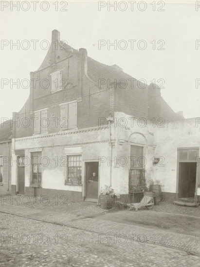 House in Amersfoort, Amersfoort 1905, photograph, title: 'Amersfoort 1905', name: 'H. Bickhoff Amsterdam', photographer: Henri Bickhoff, Amsterdam, 1905, pasteboard, paper, gelatin silver print, developing gelatin silver print, height 224 mm x width 172 mm, height 355 mm x width 298 mm, civic architecture, edifices, dwellings, Amersfoort, A two-story brick building with a stepped gable and tiled roof fills the scene, showing shuttered windows on the upper level and a mix of light and dark brickwork on the lower facade; a person is visible standing in a partially open doorway on the ground floor, next to a multi-pane window with objects on the sill and a small plant below; barrels and a wooden cart lie near the corner of the building on a cobblestone street, and a second doorway and arched window are visible along the right side, with some climbing vegetation on the wall.