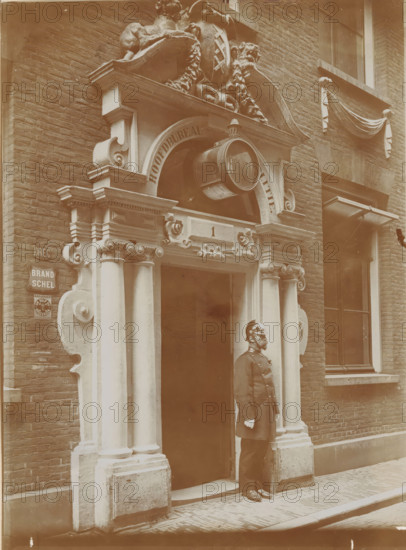 Gate of the Spinhuis, now installed in the new police station in Spinhuissteeg in Amsterdam, photograph, annotation: 'Spinhuis', Amsterdam, 1910 - 1920, photographer, paper, pasteboard, daylight gelatin silver print, height 174 mm x width 126 mm, height 290 mm x width 226 mm, policeman, constable, civic architecture, edifices, dwellings, gate, entrance, Spinhuis, A sepia-toned photograph showing an ornate stone doorway set into a brick building, featuring carved columns with decorative capitals, curved scrollwork, and an arched pediment with sculptural ornament above. A round hanging sign projects from the arch, and a carved rectangular panel sits above the entrance. A man in a dark uniform and helmet stands at the doorway, facing sideways. To the right of the entrance is a window with a draped fabric decoration above it, and to the left a small mounted plaque on the brick wall. The street in front is paved with narrow bricks and a raised curb.