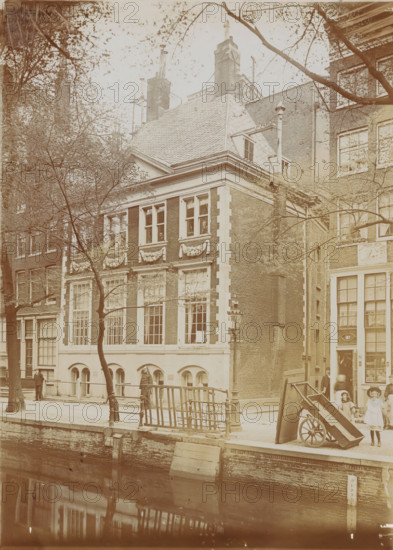 View of the Oudezijds Achterburgwal with the old Spinhuis in Amsterdam, photograph, Amsterdam, 1890 - 1920, photographer, paper, pasteboard, daylight gelatin silver print, height 173 mm x width 125 mm, height 290 mm x width 225 mm, street (city(-scape) with figures, staffage), civic architecture, edifices, dwellings, Spinhuis, A sepia-toned street scene showing a tall brick building with large multi-pane windows and decorative stonework beneath the upper windows, a steep tiled roof with dormer windows and several tall chimneys, and an adjacent building with similar windows. Trees with bare branches partially obscure the facades. A canal runs in the foreground with a stone edge and reflected shapes in the water. A railing lines the sidewalk above the canal and a lamppost stands near the corner of the building. Two children stand near a tipped wooden cart with large wheels on the pavement and a few adults stand or walk along the sidewalk near the building entrances.