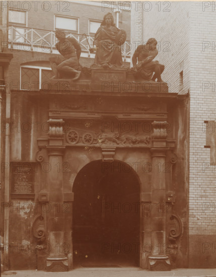 View of the Rasphuis Gate on the Heiligenweg in Amsterdam, photograph, annotation: 'Gate Heiligenweg Amsterdam, Amsterdam', name: 'Margaretha van Parma', Amsterdam, 1890 - 1910, photographer, paper, pasteboard, daylight collodion silver print, height 214 mm x width 161 mm, height 325 mm x width 235 mm, gate, entrance, Heiligeweg, A stone archway set into a building façade with decorative columns and ornate scrollwork surrounds a dark recessed entrance, above which sits a sculpted frieze featuring carved relief designs and wheel motifs, and higher still three large statues are positioned on a ledge: a seated robed female figure holding a round object flanked by two muscular male figures in reclining poses, all framed by adjacent brick and tiled walls and windows above.