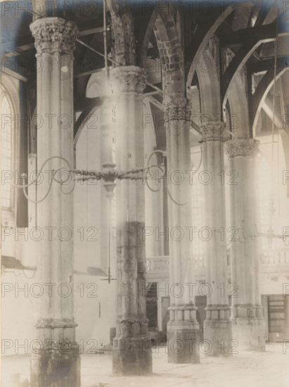 Interior of the Nieuwezijds Chapel in Amsterdam, photograph, annotation: 'Amsterdam, NZ Chapel, Interior, Demolished, Aug: 1908', photographer: Albert Adriaansz (possibly), 1908, paper, pasteboard, gelatin silver print, developed gelatin silver print, height 212 mm x width 158 mm, height 325 mm x width 235 mm, interior of church, Amsterdam, Nieuwezijds Chapel, Tall stone columns with ornate carved capitals rise through a space with pointed arches and exposed wooden beams, pale light filtering through tall arched windows. A metal chandelier with decorative curved arms hangs across the foreground, its silhouette partially obscuring the columns. The stone bases of the columns show wear and textural variation, and vertical shafts extend upward to intersect with ribbed arches and timber trusses. In the background a balcony with balustrade and a stair are visible between the columns, and the floor appears pale and worn. The overall scene is monochrome and softly lit, with areas of brighter l...