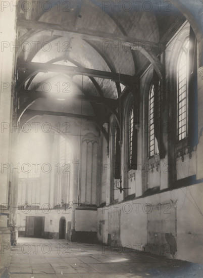Interior of the Nieuwezijds Chapel in Amsterdam, photograph, annotation: 'Amsterdam, NZ Chapel, View into the side aisle, demolished Aug. 1908', photographer: Albert Adriaansz (possibly), Amsterdam, 1908, paper, pasteboard, gelatin silver print, developed gelatin silver print, height 216 mm x width 156 mm, height 325 mm x width 235 mm, interior of church, Nieuwezijds Chapel, A high vaulted interior with wooden arched beams crossing the ceiling and large tall arched windows along the right wall, light streaming through one window and creating bright flares. The walls show worn plaster and decorative stone corbels beneath the windows, with a ledge running along the right side. In the left and central area there are tall columns and slender arches leading toward a distant wall with more arched openings and a pair of wooden doors; one doorway is topped by a small rounded arch. Suspended metal chandeliers hang from the ceiling by long rods, and thin cables or wires descend along the walls. ...