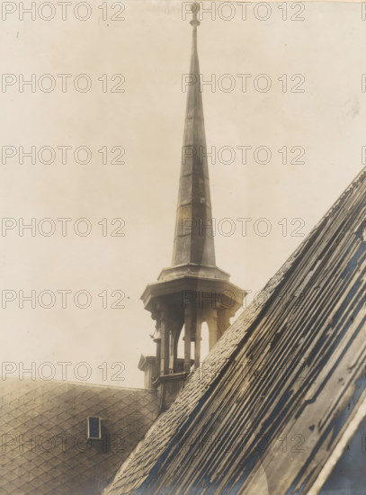 Tower of the Nieuwezijds Chapel in Amsterdam, photograph, annotation: 'Amsterdam, NZ Chapel, small tower, demolished 1908', photographer: Albert Adriaansz (possibly), Amsterdam, 1908, paper, pasteboard, gelatin silver print, developed gelatin silver print, height 212 mm x width 157 mm, height 323 mm x width 236 mm, parts of church exterior and annexes: tower - QQ - small church, chapel, Nieuwezijds Chapel, A tall, narrow spire rises from a stone base with an open, columned lantern beneath it, the spire showing segmented panels with weathered surfaces and a rounded finial at the top; adjacent sloped roofs with overlapping tiles occupy the foreground, one roof showing a small rectangular vent set into patterned shingles, and the image overall has a muted, aged tone with subtle stains and specks across the sky area.