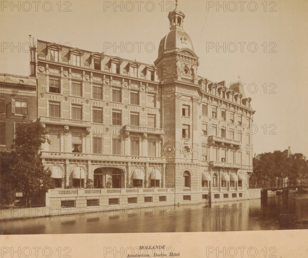 View of the Doelen Hotel in Amsterdam, photograph, inscription: 'Hollande, Amsterdam. Doelen Hôtel', annotation: 'Amsterdam', Amsterdam, 1883 - 1900, photographer, paper, pasteboard, albumen print, height 271 mm x width 280 mm, height 263 mm x width 332 mm, hotel, hostelry, inn, Doelenhotel, A wide sepia-toned photograph of a large ornate building along a canal, showing multiple stories with rows of windows, decorative stonework, and mansard-style dormer windows on the roof; the central section features a prominent tower with a domed roof, clock face, sculpted details, and a small open belfry at the top; the ground level has arched openings with striped awnings, columns, and a balustraded terrace with potted plants; the canal in the foreground reflects the building facade; trees and a fence appear at the sides, and a distant rooftop silhouette is visible on the right.