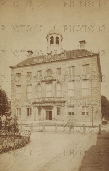Enkhuizen Town Hall, photograph, cabinet photograph, annotation: 'VI, 1 c', inscription: 'Enkhuizen. Hotel de ville', Enkhuizen, 1870 - 1900, photographer, paper, pasteboard, albumen print, albumendruk, height 181 mm x width 115 mm, height 194 mm x width 123 mm, town hall, Stadhuis (Enkhuizen), A large stone building with a symmetrical façade rises from a paved approach, featuring evenly spaced rectangular windows and decorative stonework around a central entrance with steps and a recessed doorway framed by columns and an arched transom. Above the entrance is an ornate balcony area with sculpted figures and a circular window set into the wall, and a small dome-like cupola sits atop the roof with open arches. Chimney-like structures project from the roof on either side, and a low wooden fence runs across the foreground beside a dirt path. A lamppost stands near the building's right side, a cart is positioned near the entrance, and trees and trimmed hedging appear at the left and right e...