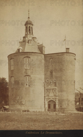 View of the Drommedaris in Enkhuizen, photograph, cabinet photograph, annotation: 'VI, 1a', inscription: 'Enkhuizen Le Drommédaire', Enkhuizen, 1870 - 1900, photographer, paper, pasteboard, albumen print, albumendruk, height 179 mm x width 115 mm, height 194 mm x width 123 mm, harbour, Drommedaris (Enkhuizen), A large round brick building with two cylindrical towers connected by a curved wall occupies the image, showing multiple tall rectangular windows and a decorative arched entrance with sculpted stonework; a steep slate roof rises to a cupola with arched openings and a bell visible inside, topped by a finial and weather vane; a flagpole stands on the roof near a small chimney; a grassy foreground with short posts and a fence runs across the base of the building, and partial neighboring structures and trees are visible at the sides.
