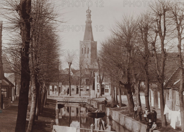 View of Medemblik with the tower of the Reformed Church, photograph, inscription: 'B. Reekers, Photographer, Enkhuizen', annotation: 'View of the tower of the Reformed Church in Medemblik, Photo B. Reekers', annotation: 'N:6, 13 1, 2 wide', photographer: Barend Reekers, Medemblik, 1890 - 1920, paper, pasteboard, daylight collodion silver print, height 126 mm x width 173 mm, height 141 mm x width 186 mm, canals, waters (in city), church (exterior), street (city(-scape) with figures, staffage), A tree-lined street runs beside a narrow canal with small rowboats moored along the stone edge, bare branches of the trees forming a lattice against the sky; buildings with pitched roofs and multiple windows face the water, and a tall church tower with a steep spire and an ornate top rises above the rooftops; people stand and sit along the canal bank and near the buildings, and a line of laundry hangs between trees, all reflected in the still surface of the canal.