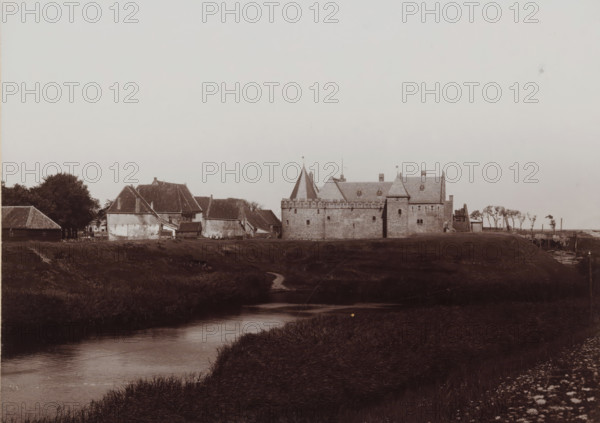 View of the former Koggenhuis in Medemblik, photograph, annotation: 'Medemblik, Former Koggenhuis in Medemblik', inscription: 'B. Reekers, Photographer, Enkhuizen', photographer: Barend Reekers, Medemblik, 1890 - 1915, paper, pasteboard, daylight collodion silver print, height 121 mm x width 167 mm, height 173 mm x width 126 mm, urban housing, Radboud Castle, A wide view of a low-lying landscape with a meandering waterway in the foreground and grassy banks leading toward a row of buildings on the far bank, including masonry structures with battlemented walls and steeply pitched roofs, a conical tower with a small flag, and adjacent cottages with tiled roofs and weathered exterior walls, clusters of trees near the buildings, a narrow dirt path running from the bank down to the water, and a strip of open sky above.