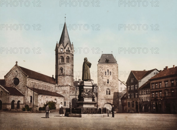 View of the Karlsplatz in Eisenach with the statue of Martin Luther, Eisenach, Karlsplatz mit Lutherdenkmal, photomechanical print, number: '6448.', cliché maker: Photochrom Zürich, Zürich (city), 1889 - c. 1920, photographer, Thüringen, pasteboard, paper, photochrom, height 188 mm x width 248 mm, Person, Martin Luther, monument, statue, square, circus, Karlsplatz, A stone plaza scene with a tall statue of a robed figure on an ornate pedestal surrounded by a low decorative fence, a large church building with a tall pointed bell tower and visible clock face, an adjacent square stone tower with small arched windows, and rows of multi-story buildings with pitched roofs; a few people stand near the statue and along the plaza, and the sky above is pale blue.