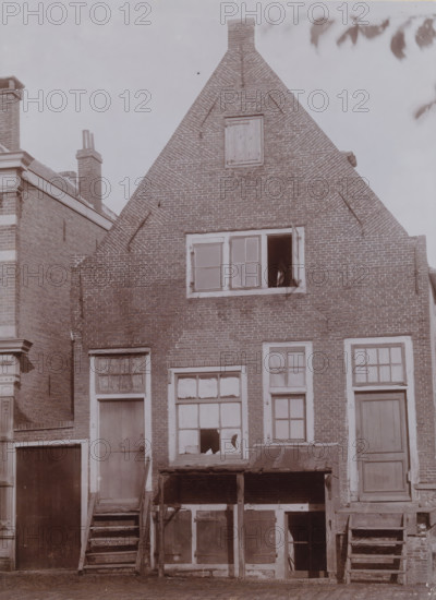 Façade of Buitenveer 141 in Weesp, photograph, annotation: 'Buitenveer 141, demolished Nov. 1911, Weesp', Weesp, photographer: AJM Mulder (possibly), 1910 - 1911-11, Attributed to, paper, pasteboard, daylight collodion silver print, height 210 mm x width 154 mm, urban housing, Buitenveer, A brick gabled building with a steep triangular roof and a small boarded opening near the top stands centered in the image, showing patterned brickwork and a chimney at the apex; a rectangular window with one open pane is visible below the midpoint, and beneath that are several framed windows and doors with peeling paint and broken glass, some set above wooden steps and small platforms supported by posts; adjoining brick structures with chimneys and decorative trim appear at the left and right edges, and tree leaves are visible at the top right against a pale sky.