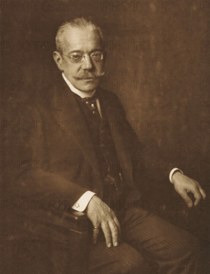 Portrait of Dr. James Simon, photomechanical print, signature: 'H. James Simon', inscription: 'PHOT. DÜRKOPP', photographer: Rudolf Dürkopp, Berlin, cliché maker: anonymous, 1895 - 1910, pasteboard, paper, photoaquatint, height 291 mm x width 223 mm, height 405 mm x width 290 mm, Person, James Simon, historical persons, adult man, A sepia-toned portrait of an older man seated against a plain backdrop, wearing round spectacles and a neatly groomed mustache, dressed in a dark suit with a white shirt collar and a visible tie, his hands resting on his lap and the arm of a chair, one hand partly gloved by a visible cuff, the image showing textured grain and soft lighting.