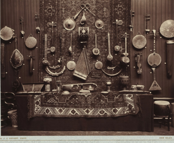 Musical instruments from the Daniël François Scheurleer collection, exhibited in Pulchri Studio in 1893, photomechanical print, inscription: 'A. vd GRIENT, PHOT. - THE HAGUE.', photographer: Adrianus van der Grient, Pulchri Studio, 1893, The Hague, pasteboard, paper, Woodburytype, Woodburytypie, height 220 mm x width 280 mm, Person, Daniël François Lunsingh Scheurleer, musical instruments, group of musical instruments, exhibition, art, musical instrument, A large patterned rug hangs vertically as a backdrop with a variety of stringed and percussion instruments arranged on and around it, including lutes, harps, bowed instruments, round frame drums, cymbals, and carved wooden pieces, some suspended from pegs and others leaning against the rug; a tapestry with geometric motifs covers a table in front of the display, on which rests smaller vessels, bowls, a few additional hand drums, and a curved wooden horn; the scene is sepia-toned and shows wood-paneled walls on either side of the centr...