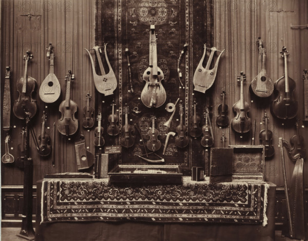 Musical instruments from the Daniël François Scheurleer collection, exhibited in Pulchri Studio in 1893, photomechanical print, inscription: 'A. vd GRIENT, PHOT. - THE HAGUE.', photographer: Adrianus van der Grient, Pulchri Studio, 1893, The Hague, pasteboard, paper, Woodburytype, Woodburytypie, height 221 mm x width 280 mm, Person, Daniël François Lunsingh Scheurleer, exhibition, art, string instruments (plucked), musical instrument, A wall covered with a patterned hanging and vertical wooden panels displays a variety of stringed instruments suspended at different heights, including several violins, cellos, a double bass, lyres, lutes, and other plucked and bowed instruments with visible tuning pegs and carved bodies; a large carved harp stands to the right edge. Below the instruments a table draped with an ornate patterned textile holds open cases, a small box, and other accessories, while a long rug with repeating geometric motifs hangs centrally behind the instruments, showing worn...