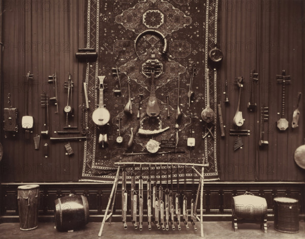 Musical instruments from the Daniël François Scheurleer collection, exhibited in Pulchri Studio in 1893, photomechanical print, inscription: 'A. vd GRIENT, PHOT. - THE HAGUE.', photographer: Adrianus van der Grient, Pulchri Studio, 1893, The Hague, pasteboard, paper, Woodburytype, Woodburytypie, height 220 mm x width 280 mm, Person, Daniël François Lunsingh Scheurleer, musical instruments, group of musical instruments, exhibition, art, musical instrument, A large patterned rug hangs centrally on a vertical paneled wall, with a variety of stringed and percussion instruments mounted around and on it; several long-necked lutes and rounded-bodied instruments hang from the rug and wall, some with carved necks and pegboxes, and smaller hand-held instruments with curved shapes are interspersed; a pair of crescent-shaped objects and a circular drumhead are visible on the rug; numerous slender pipes arranged in a wooden frame stand in front of the rug, their open ends facing outward; drums of d...