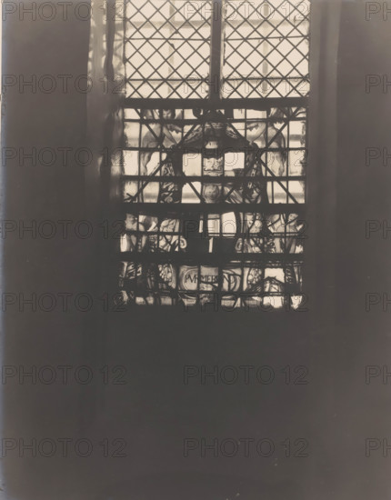 Painted window in the Castle Chapel Egmond in Egmond aan den Hoef, photograph, annotation: 'Egmond a, d Hoef, stained glass window of Purmerend 1634 in the south facade of the Dutch Reformed Church, Feb 1907', annotation: 'stained glass window of Purmerend in the south facade of the Dutch Reformed Church, Feb. 1907, Egmond a, d Hoef', Castle Chapel (Egmond-aan-den-Hoef), 1907, photographer, Egmond aan den Hoef, Attributed to, paper, pasteboard, gelatin silver print, height 205 mm x width 163 mm, interior of church, church window, e.g. rose window, A tall window with diamond-patterned leaded panes fills the upper center of the image, showing a grid of lighter glass panels framed by darker mullions; behind the panes there are decorative painted or stained sections with figurative shapes and ornamental motifs visible through the lower portion of the window, and darker vertical wall surfaces flank the window on both sides, while the lower two-thirds of the image is dominated by a large dar...